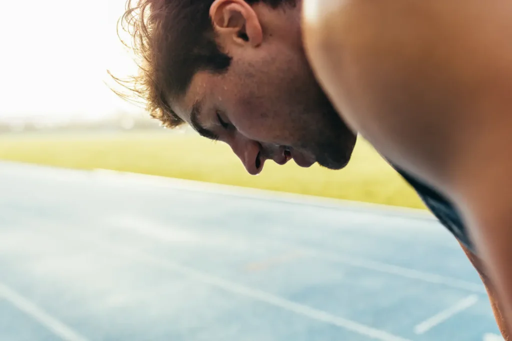 Close-up of a man looking down with an expression of exertion or fatigue. He is on a track with blurred green grass and blue track markings in the background. Warm lighting suggests it is sunrise or sunset, adding a golden atmosphere.
