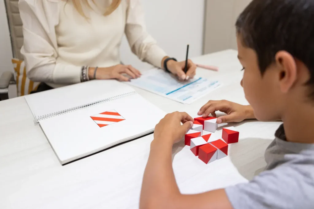 A child interacts with red and white geometric blocks on a table, possibly part of a task or game. An adult woman in the background is seated at the table, writing on a piece of paper, seemingly supervising or assisting. In front of the child is a notebook or sketchbook featuring designs with red and white triangular patterns.