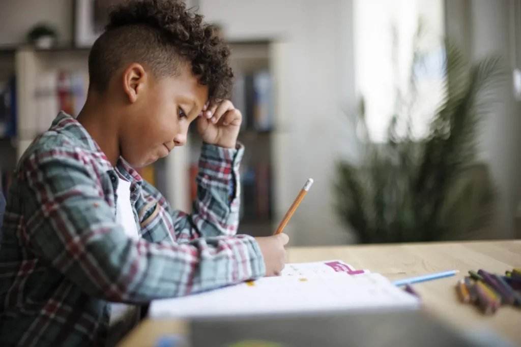 A child with curly hair and wearing a plaid shirt sits at a table, deeply focused on writing or drawing on a piece of paper. Their head rests on one hand in thought, surrounded by scattered colored pencils. The background is blurred, showing shelves and plants in a cozy indoor setting.