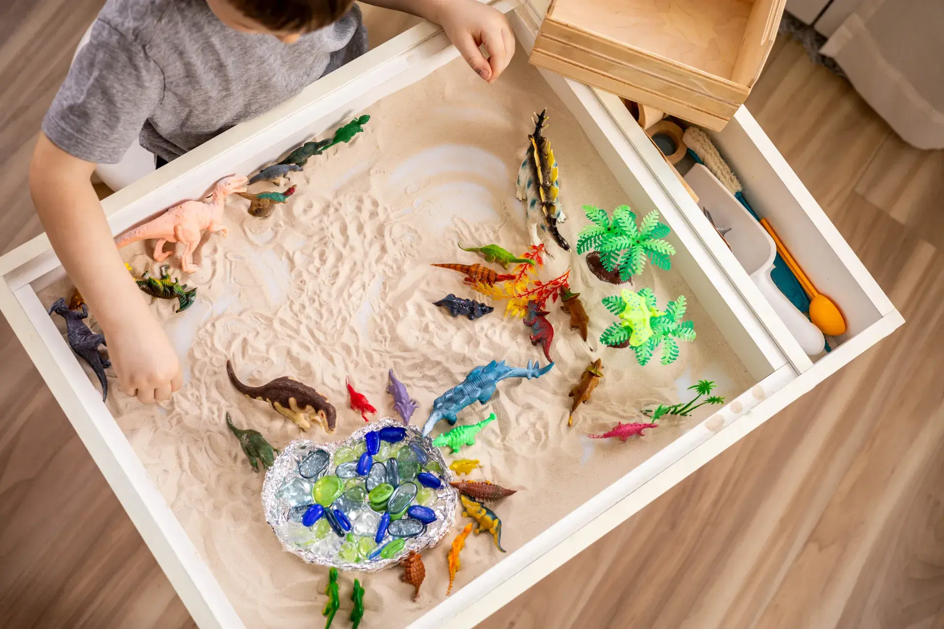 A sensory play area featuring a sand table where a child is playing with toy dinosaurs and small plastic plants. The sand includes a foil dish filled with colorful stones resembling water, encouraging imaginative play. Additional tools and items are visible in a drawer above the sand table.
