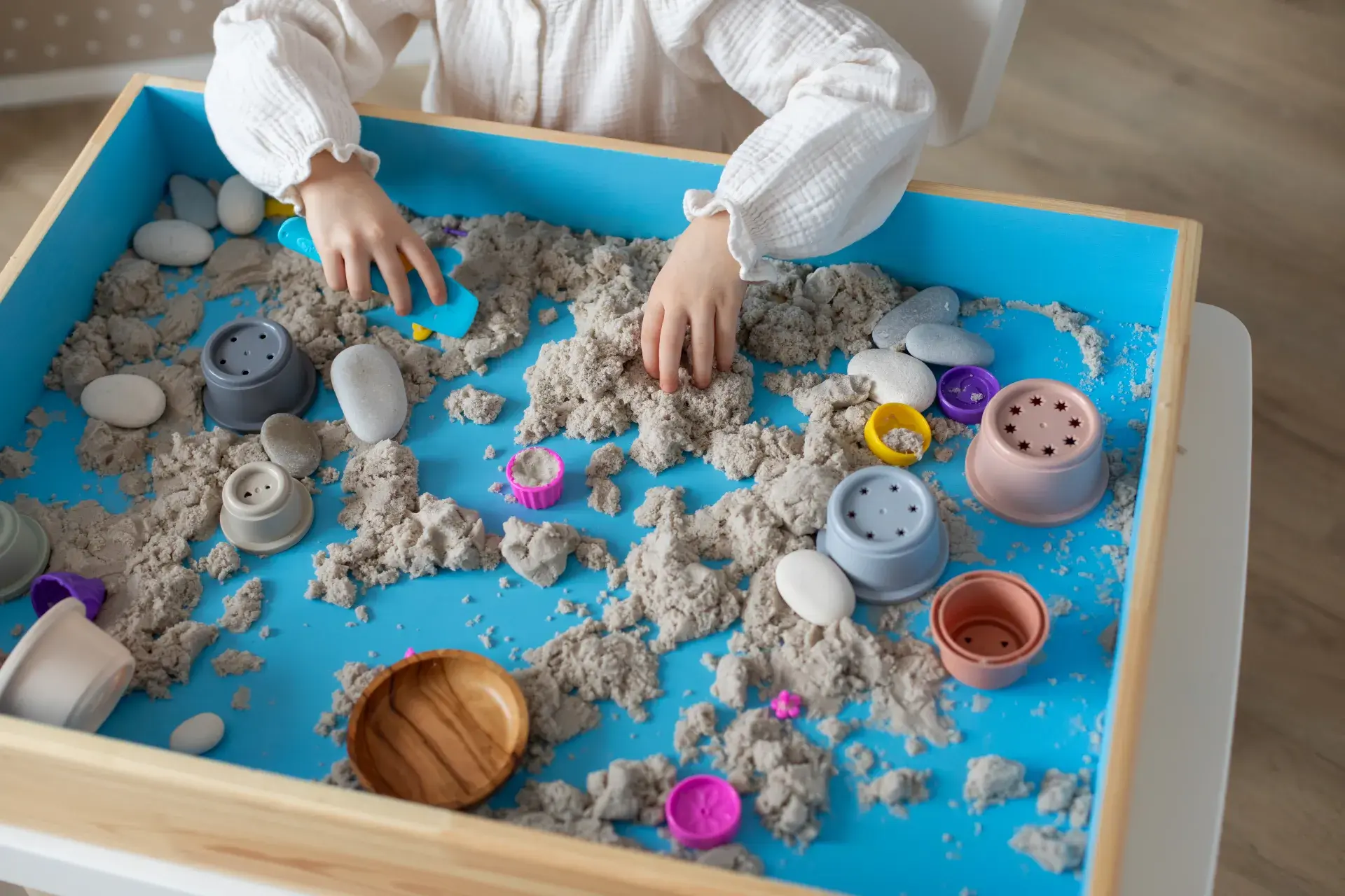A sensory play setup featuring a blue tray filled with light brown, sand-like material and small smooth stones. Colorful plastic molds, tools, and small containers are scattered throughout. A child's hands are actively engaging with the materials, highlighting a playful and creative tactile exploration activity.