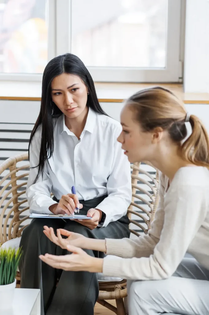 Two women are having a conversation in a cozy, well-lit room. One woman, seated in a chair and wearing a white shirt with dark pants, is taking notes on a clipboard. The other woman, dressed in a light sweater and seated opposite, has her hands raised as she speaks or expresses herself. The setting suggests a professional consultation or counseling session.