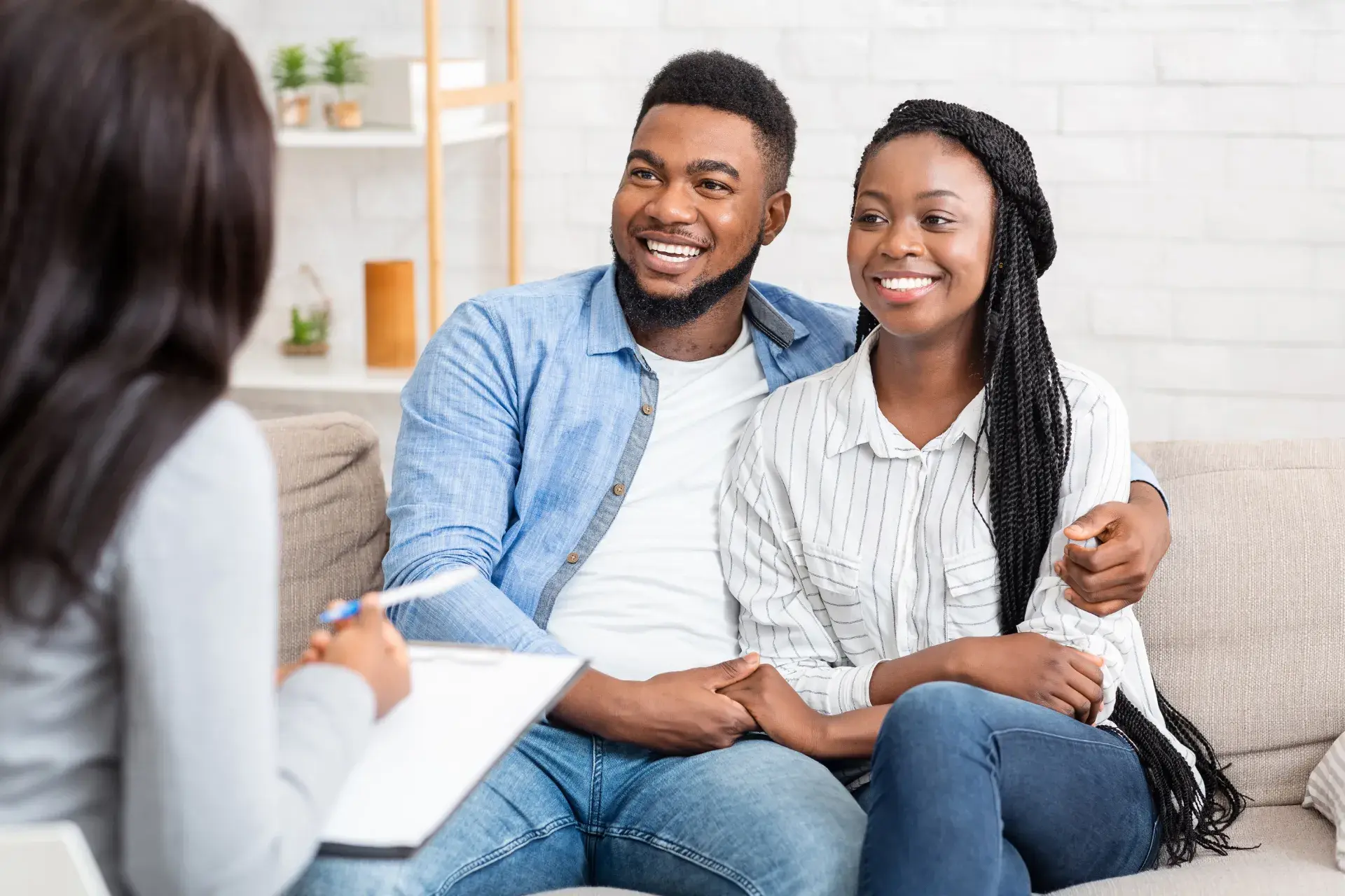 A couple sitting on a couch, looking happy and engaged, while a therapist or counselor sits across from them holding a clipboard and listening attentively. The setting is relaxed with plants and decorative items in the background.
