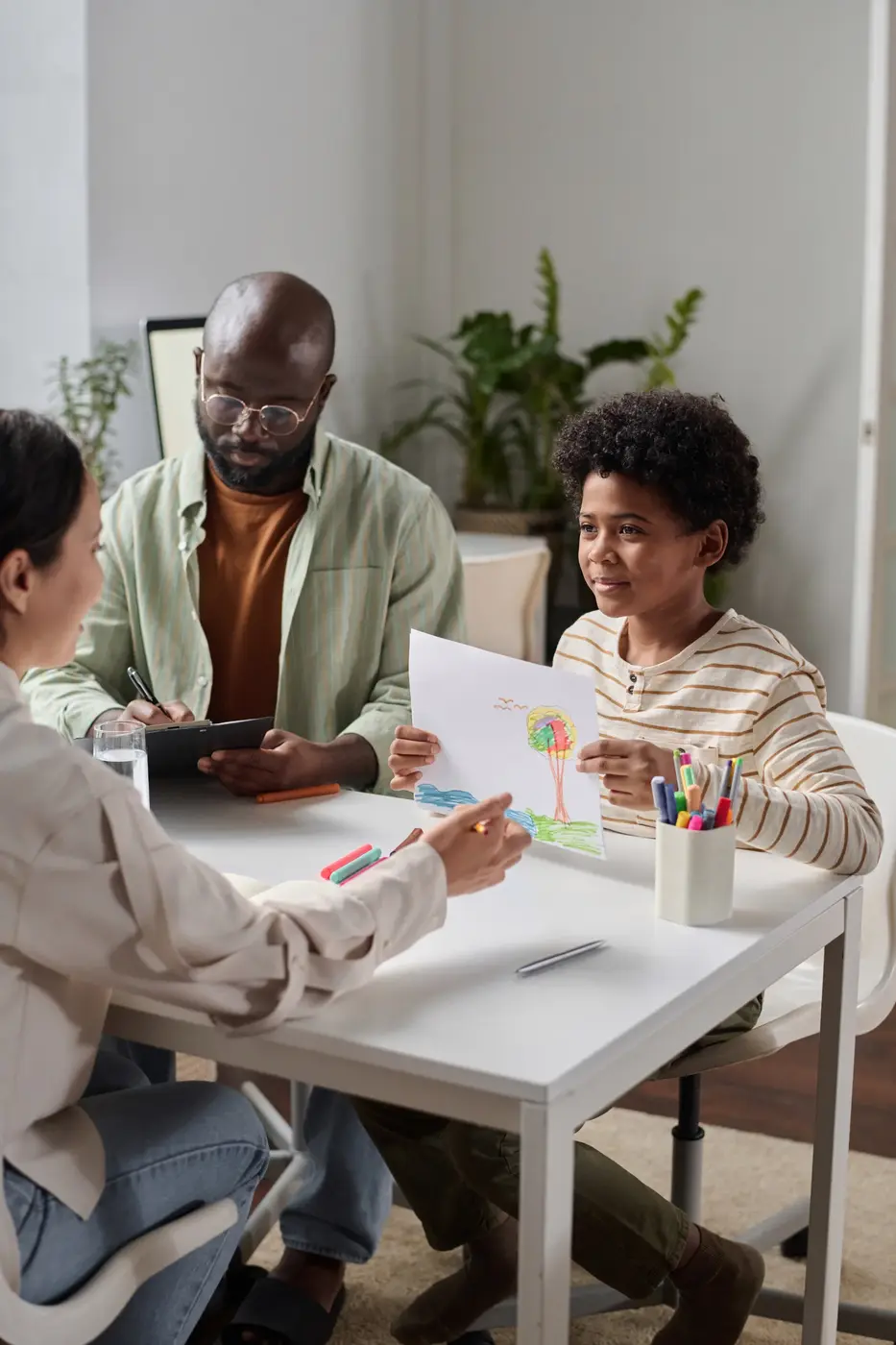 A child holds up a colorful drawing of a tree and clouds while smiling at a table. An adult, possibly a parent or guardian, engages with the child. Another adult sits nearby looking at a device. The table has markers, a cup of colored pencils, and a glass of water. The room is bright and modern, with plants in the background.