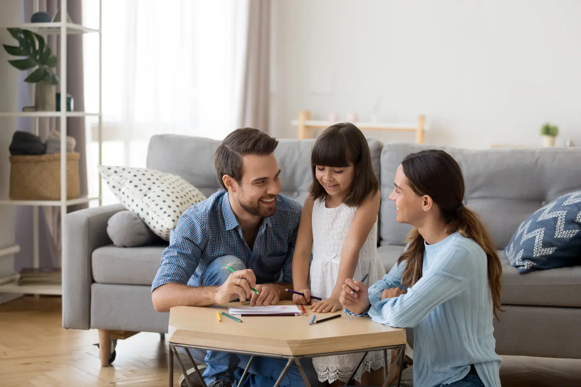 Parents and their young daughter talking and drawing together at a small table in a bright living room