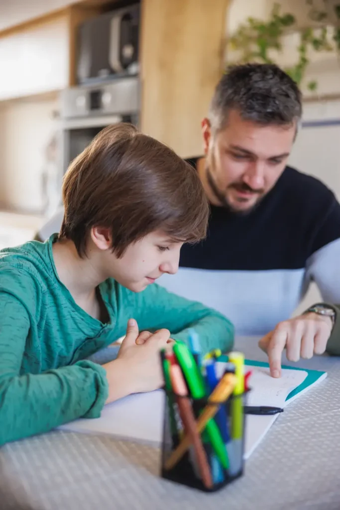 A young boy with short hair sits at a table next to a man in a home setting. The boy is looking down, appearing focused, while the man points at a piece of paper, seemingly explaining something. Also on the table are a container filled with colorful pens or markers and some paper, suggesting a learning or creative activity.