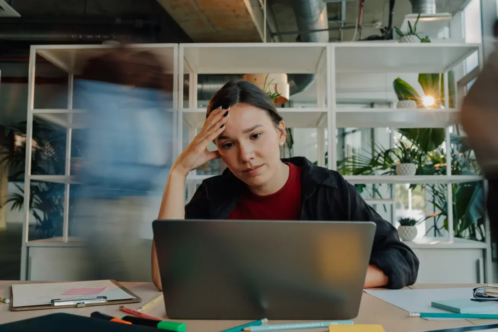 A woman sitting in an office setting, looking stressed or concerned while working on a laptop. She has her hand on her forehead and is deeply focused on the screen. In the background, a blurred figure suggests movement, and there are indoor plants, pens, and papers on the table. The scene conveys a sense of urgency or pressure.
