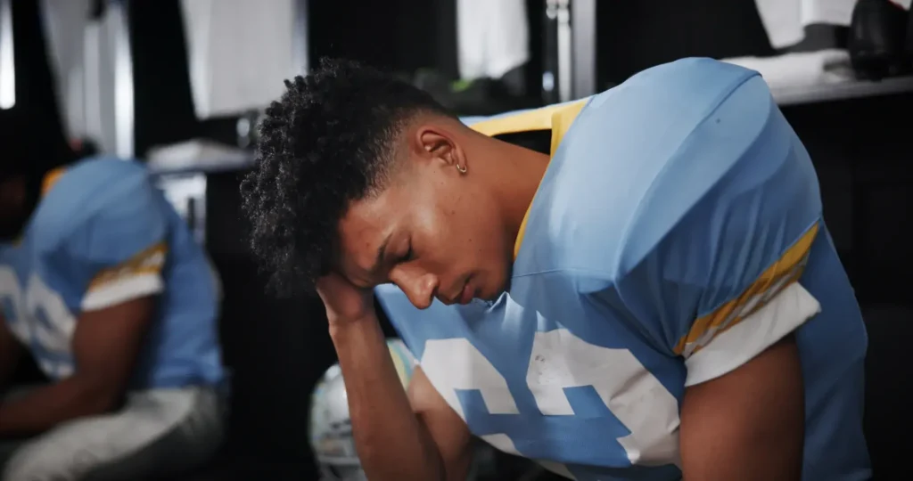 A football player wearing a jersey with the number '44' sits in a locker room with a contemplative expression, possibly reflecting or feeling stressed. Another player, partially visible in the background, is also in a football uniform, suggesting a pre-game or post-game moment.