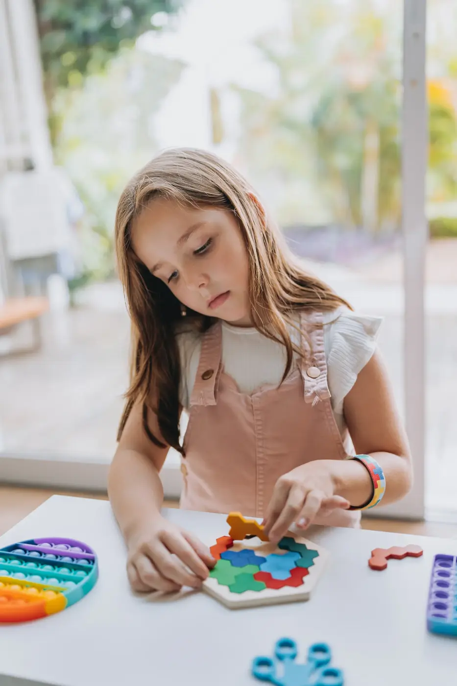 A young girl sits at a table, focused on solving a colorful puzzle with a hexagonal wooden board. Various puzzle pieces are scattered around her, along with a multi-colored silicone sensory toy. The background features greenery, indicating a bright and cheerful indoor environment.
