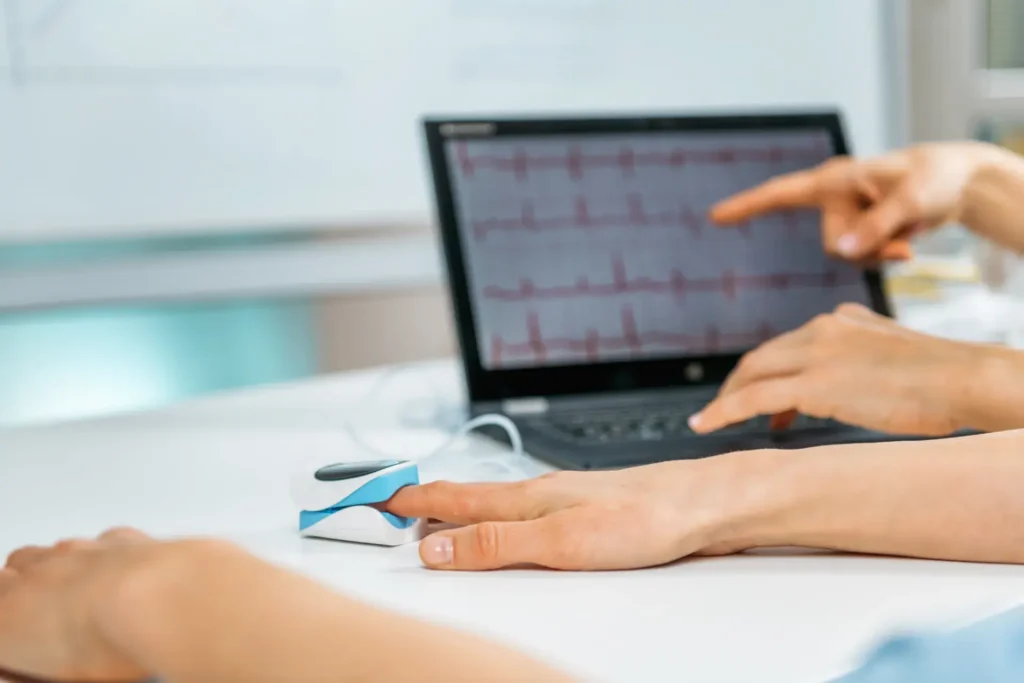 A laptop displays a graph with peaks and troughs, resembling medical data such as an ECG. One hand is placed on a small device, possibly measuring vital signs, while another hand points at the laptop screen, indicating a discussion or analysis of the data in a clinical or research environment.
