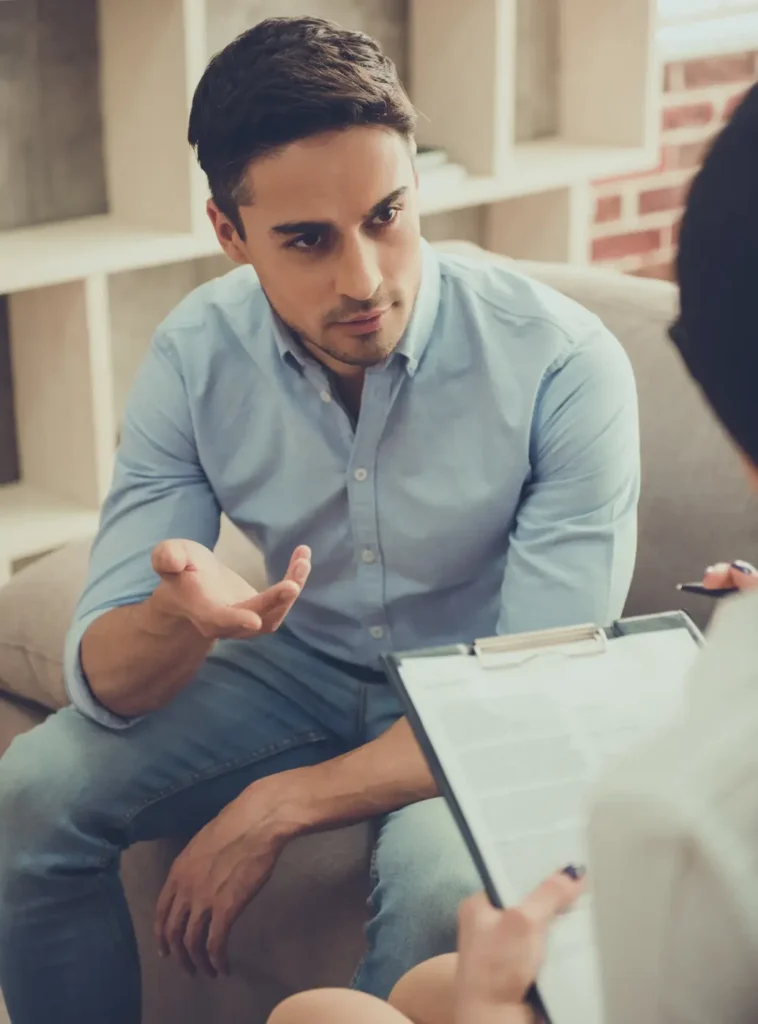 A man sits on a couch in a relaxed setting, appearing to engage in a serious conversation. His expression is thoughtful, and one hand is raised as if making a point. A clipboard with papers lies in front of him, suggesting he might be participating in a counseling or therapy session in an informal, comfortable office or home environment.