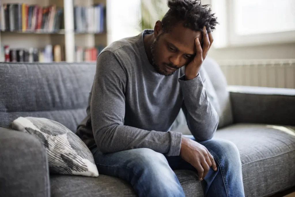 A somber man sits on a couch, wearing a gray long-sleeve shirt and jeans, with his head resting on his hand in a contemplative or distressed pose. The background has shelves with books, creating a cozy, home-like setting.