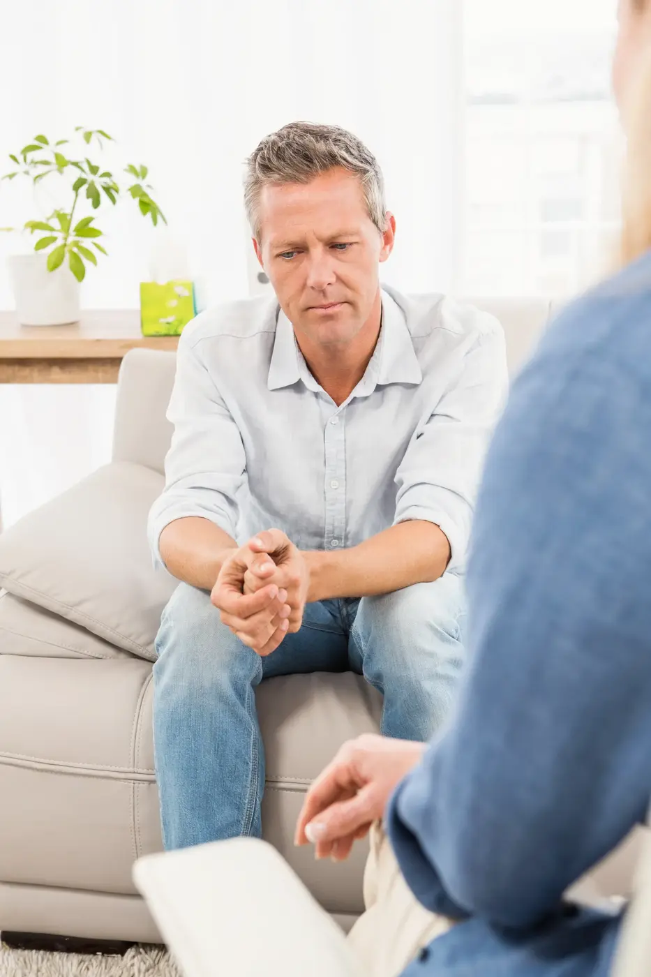 A man with short hair sits on a couch, looking serious or thoughtful. He is wearing a light-colored shirt with rolled-up sleeves and jeans. In the background, there is a plant on a shelf and a partially visible person in denim clothing facing him. The setting is a cozy indoor space, possibly an office or meeting room, suggesting a conversation or discussion is happening.