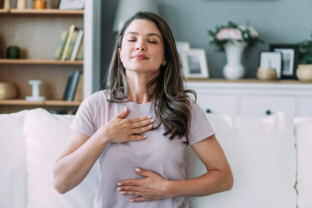 A woman sits on a couch with a calm expression, her eyes closed. One hand rests on her chest and the other on her abdomen, indicating she is practicing mindfulness or deep breathing. In the background, a shelf with books and decorative items adds to a cozy home setting.