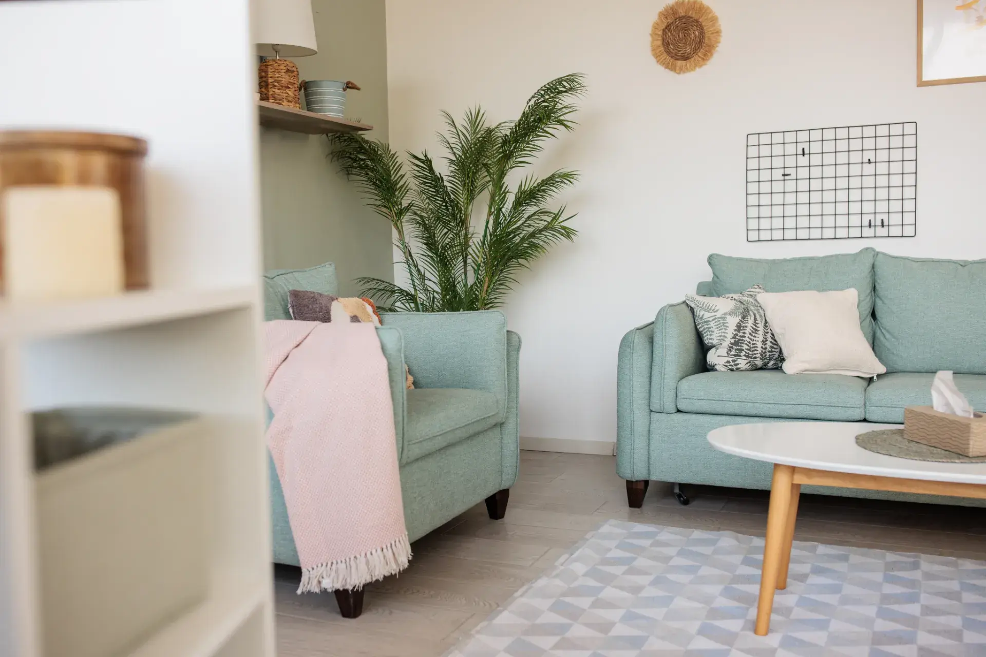 A cozy living room featuring two light blue sofas with various cushions, one patterned and one plain, and a light pink throw draped over a sofa. A potted plant adds greenery in the background. A white coffee table with wooden legs holds a decorative tray, placed on a light, geometric-patterned rug. The room is decorated with soft, neutral tones, a woven wall decoration, and a grid wall feature, with light-colored flooring completing the space.