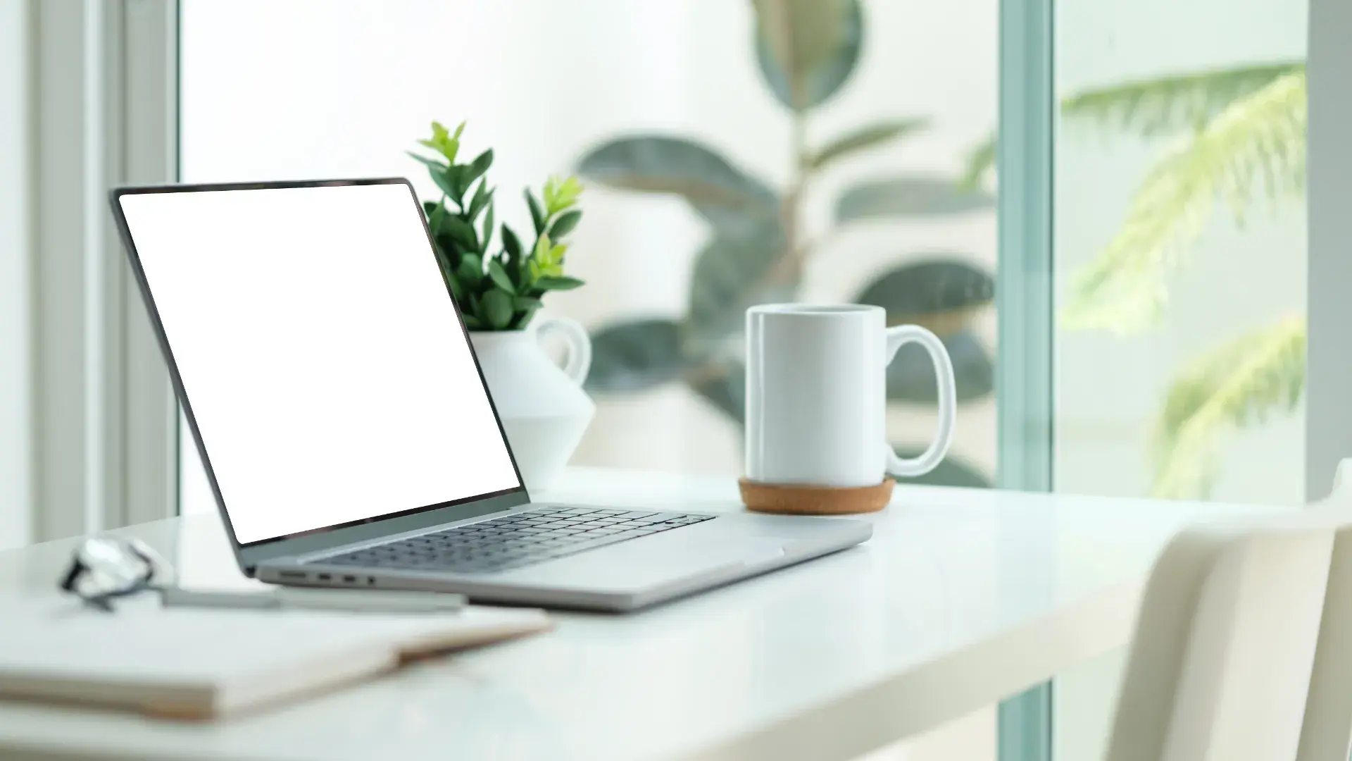A modern workspace featuring a laptop with a blank screen, a small potted plant in a white container, a white mug on a circular coaster, and a notebook placed on the desk. The area is brightly lit with natural light, suggesting proximity to a window.