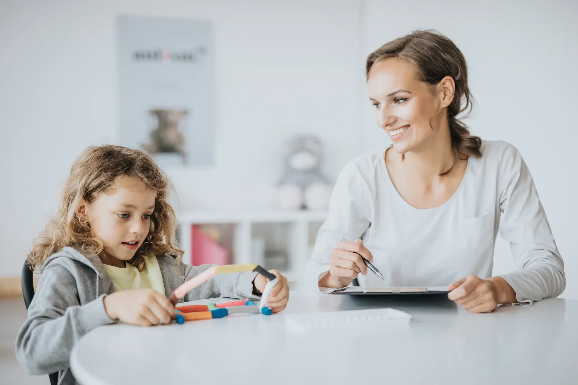 Girl building with colourful sticks at a white table while a smiling adult looks on