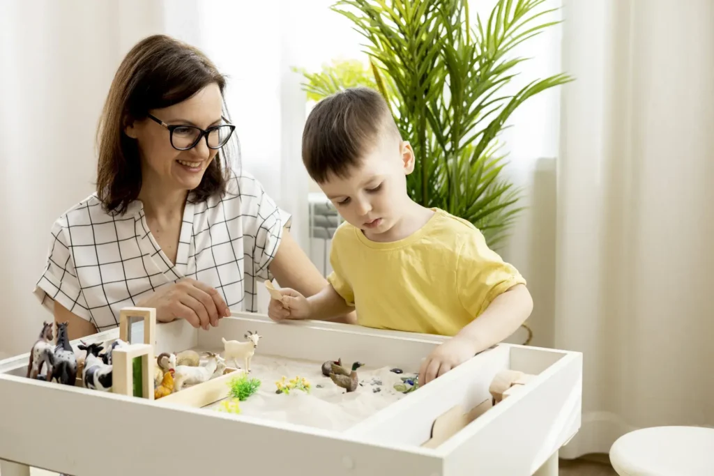 A woman wearing a checkered shirt smiles and helps a young boy in a yellow shirt as they play with toy animals and decorative elements in a sandbox-like container. The setting is bright and cheerful with a green plant visible in the background.