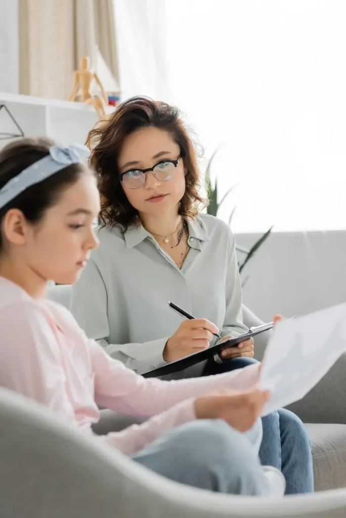 A woman and a girl are sitting together in a bright, modern environment. The woman is writing on a clipboard while the girl sits nearby, looking at a piece of paper. The background features plants and decorative items, creating a welcoming atmosphere.