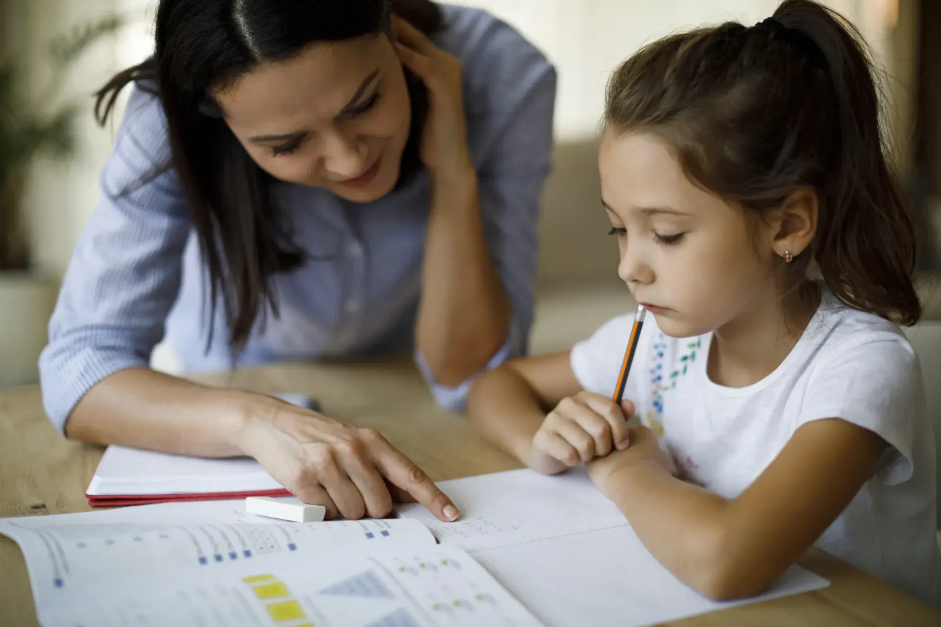 A woman and a young girl working together at a table on a learning activity. The woman is pointing at a sheet of paper on the table, while the girl, holding a pencil to her mouth, concentrates on the task. Papers with charts or graphs are spread on the table, indicating a school-related project. The setting appears cozy and bright, suggesting a home environment.