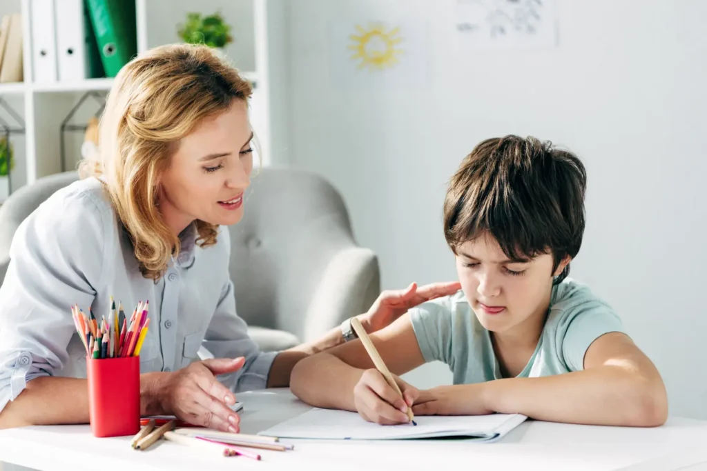 A woman leans in with a smile, assisting a young boy who is focused on writing in a notebook. A red container holding colored pencils sits on the table, and the background features a cozy setting with an armchair and wall decorations.