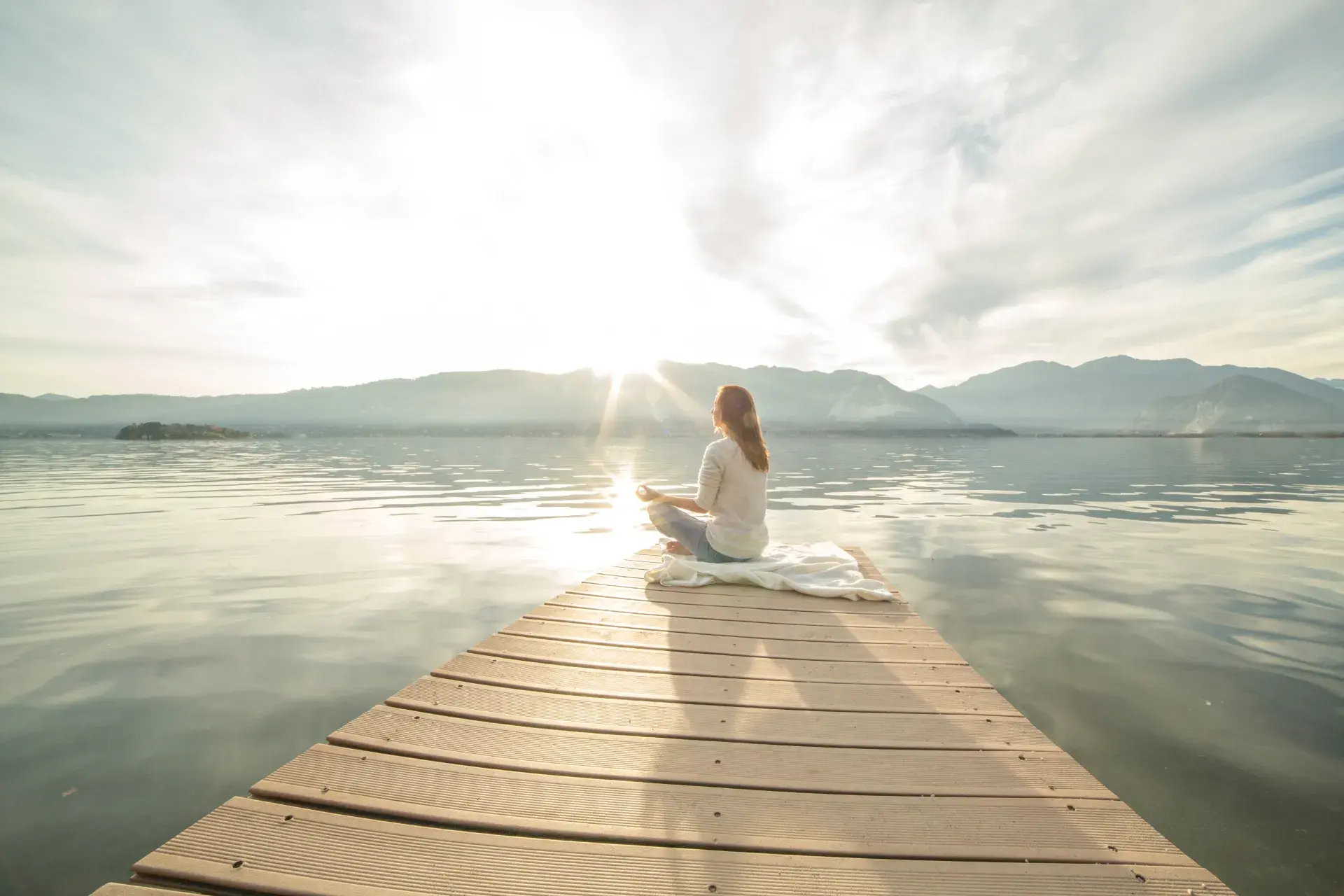 A person sits in a meditative pose on a dock extending over a calm body of water, surrounded by a scenic backdrop of mountains and a glowing sky at sunrise or sunset. They are wrapped in a blanket, exuding relaxation and mindfulness, with a tranquil and peaceful atmosphere all around.