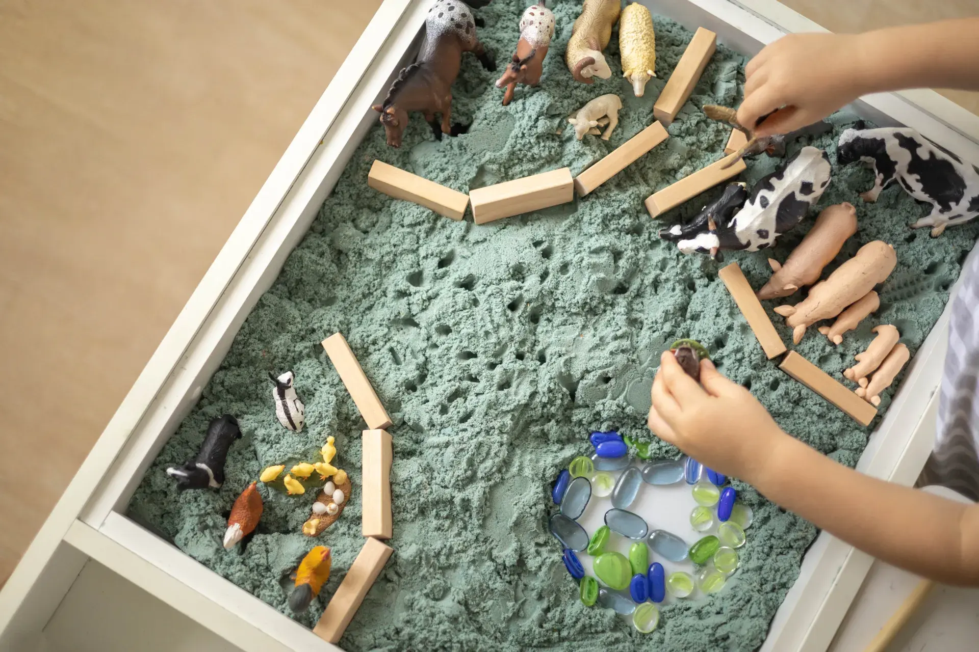 A sensory play setup featuring a tray filled with green sand, small plastic animals such as cows, pigs, and sheep, wooden blocks, and colorful beads or stones arranged in a circular pattern. A child's hand is interacting with one of the animal figures.