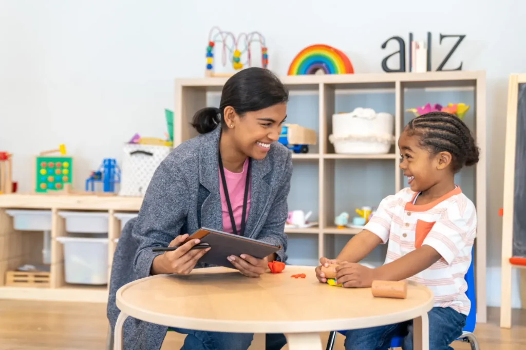 A cheerful scene of a woman and a child interacting in a playroom or classroom. The woman is holding a tablet and smiling at the child, who is seated at a circular table playing with colorful modeling clay. Shelves filled with toys and activities can be seen in the background, creating a lively and educational setting.