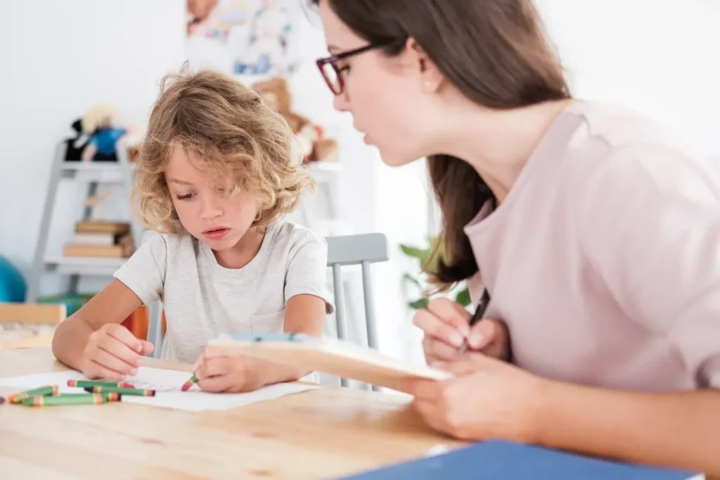 A young child with curly hair is seated at a table, intently coloring or drawing with crayons. An adult woman sits beside the child, appearing to guide or assist. The setting is a bright and cheerful space, possibly a classroom or home learning area, with colorful decor and materials visible in the background.