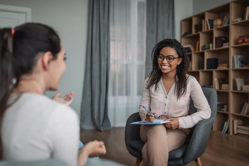 Two people converse in a relaxed, professional setting. One person, seated and smiling, takes notes while the other, slightly turned away, speaks. The background includes bookshelves and large windows with curtains, creating a calm atmosphere.