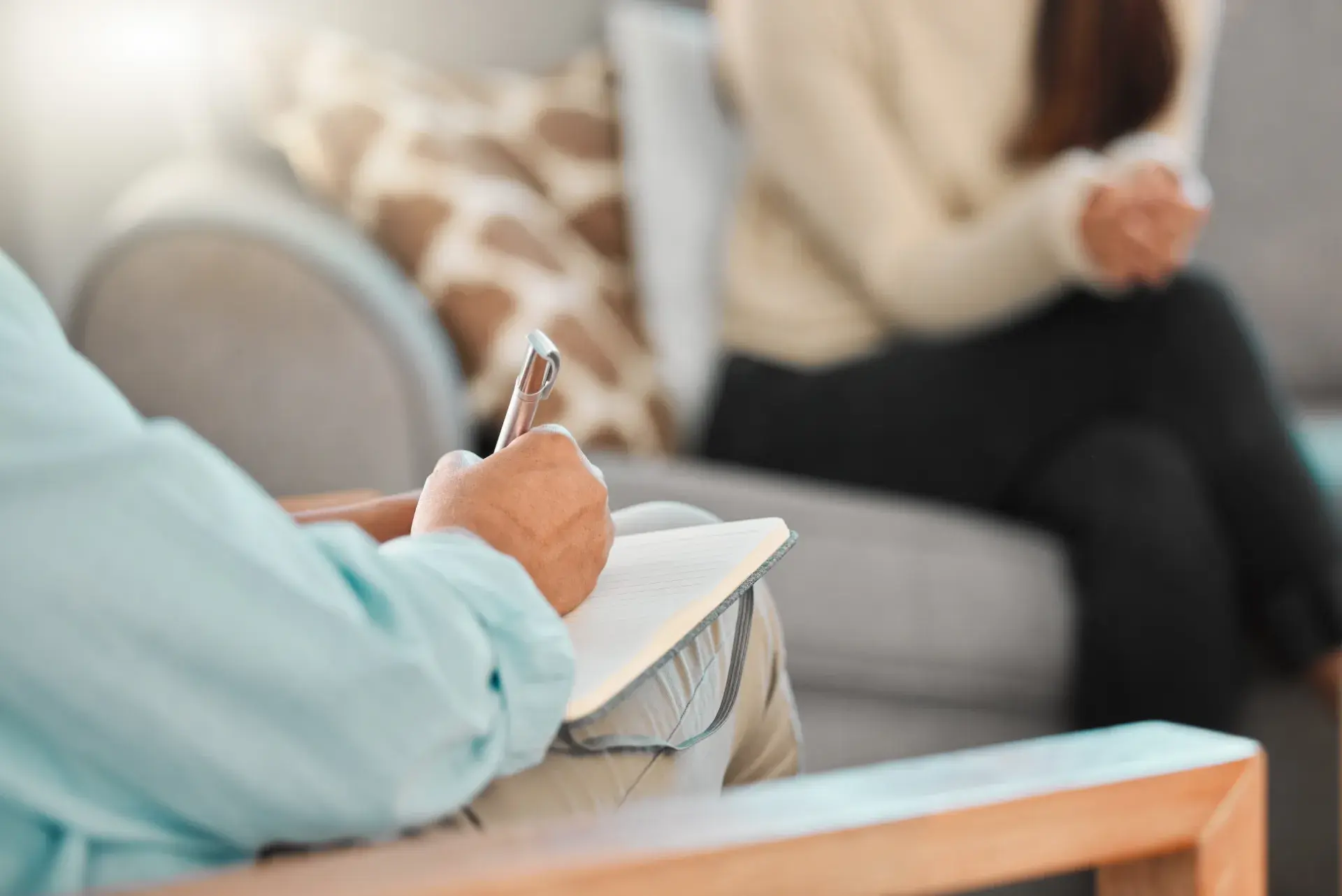 A person is sitting in a comfortable counseling setting, writing in a notebook with a pen. In the background, another individual is seated, appearing to engage in conversation. The environment includes a sofa and a decorative pillow, creating a cozy atmosphere.
