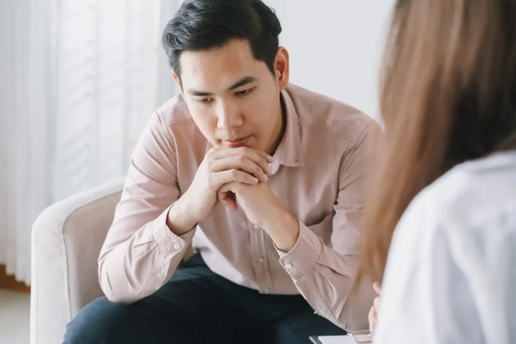 A serious man in a light-colored shirt sits on a chair with his hands clasped near his mouth, appearing contemplative or deeply focused. He seems to be engaged in a conversation or listening intently, possibly in a counseling or interview scenario. The background shows a bright, simple room with soft lighting filtering through sheer curtains, and another person's indistinct figure is partially visible.