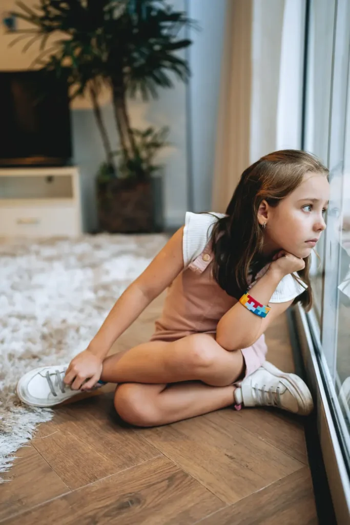 A young girl with long hair sits on the floor indoors, resting her chin on her hand as she looks out a window. She is wearing a light pink outfit and a distinctive bracelet. Behind her, there is a plant and a rug, creating a calm, reflective atmosphere.