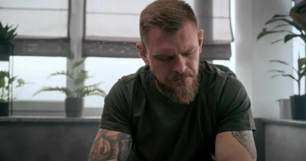 A bearded man with tattoos sits thoughtfully, looking down, surrounded by indoor plants and window coverings. The scene conveys a quiet, reflective atmosphere.