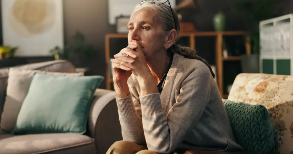An older woman with glasses resting on her head sits on a couch, her hands clasped in front of her face as she appears thoughtful or contemplative. She is wearing a sweater, and the background includes soft furnishings and decorative items, suggesting a cozy indoor setting.