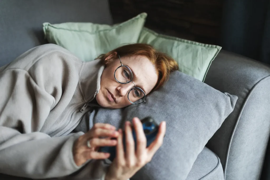 A person with red hair and glasses is lying on a couch with gray pillows, intently looking at a smartphone. They are wearing a light-colored jacket. The setting appears cozy and homely, likely indoors.